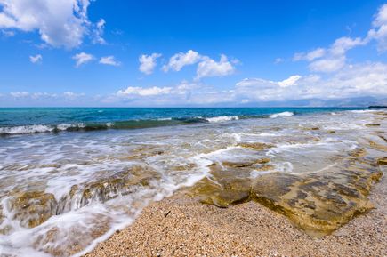 Küstenabschnitt mit flachem, felsigem Strand und sanften Wellen unter blauem Himmel mit vereinzelten Wolken