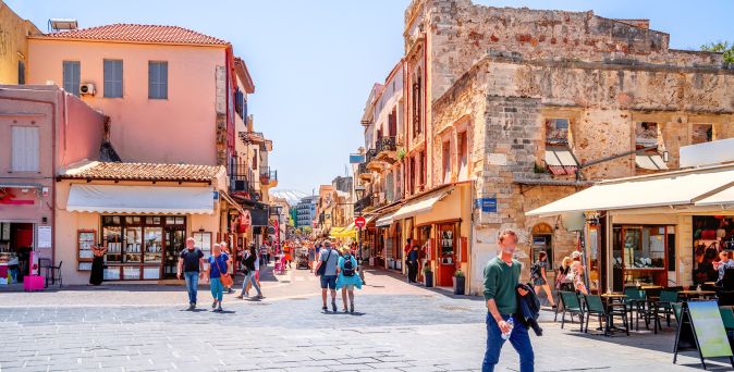 Straße in Chania auf Kreta mit historischen Gebäuden, Cafés und mehreren Passanten bei Sonnenschein.
