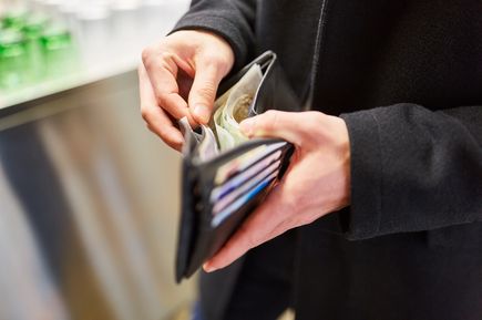 Hände einer Person in schwarzer Jacke öffnen eine schwarze Geldbörse mit sichtbaren Geldscheinen und Kartenfächern.