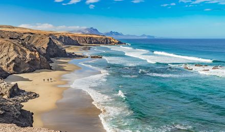 Playa del Viejo Rey auf Fuerteventura mit weitläufigem Sandstrand, rauer Felsküste, Atlantikwellen und wenigen Spaziergängern unter blauem Himmel