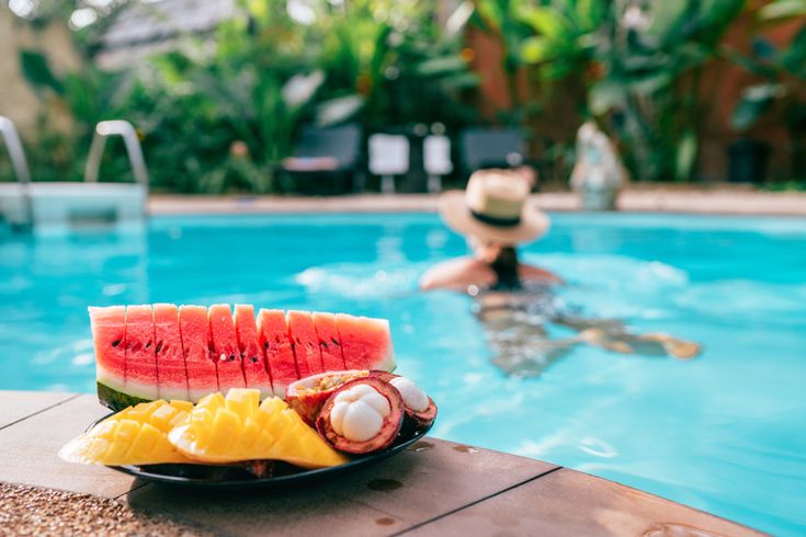 Frisches Obst auf Teller am Poolrand mit Wassermelone, Mango und exotischen Früchten, im Hintergrund Person im Swimmingpool in tropischer Urlaubsumgebung