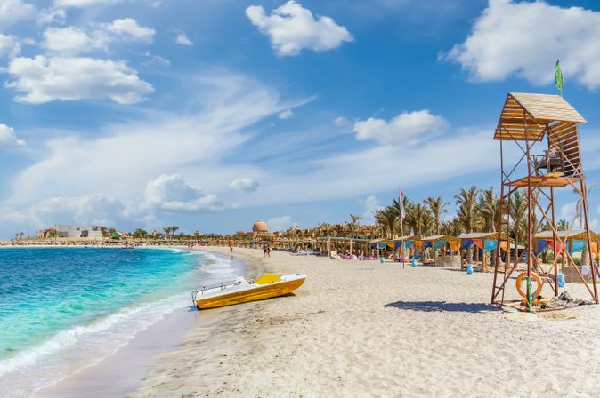 Strand mit weißem Sand, gelbem Rettungsboot am Ufer, Rettungsturm und Palmen unter blauem Himmel in Abu Dabbab, Ägypten