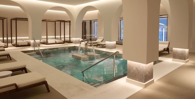 Luxuriöser Spa‑Bereich im Hotel Daios Cove mit modernem Innenpool, stilvollen Liegen, hellen Rundbögen und sanfter Ambientebeleuchtung