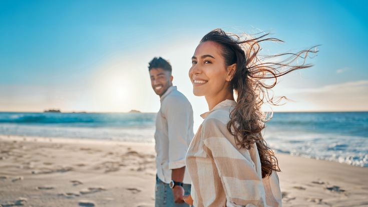 Lächelndes Paar spaziert am Strand entlang, mit Sonne und Meer im Hintergrund