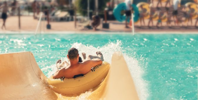 Mann und Kind rutschen gemeinsam auf einem gelben Schwimmreifen eine Wasserrutsche hinunter und landen im Pool