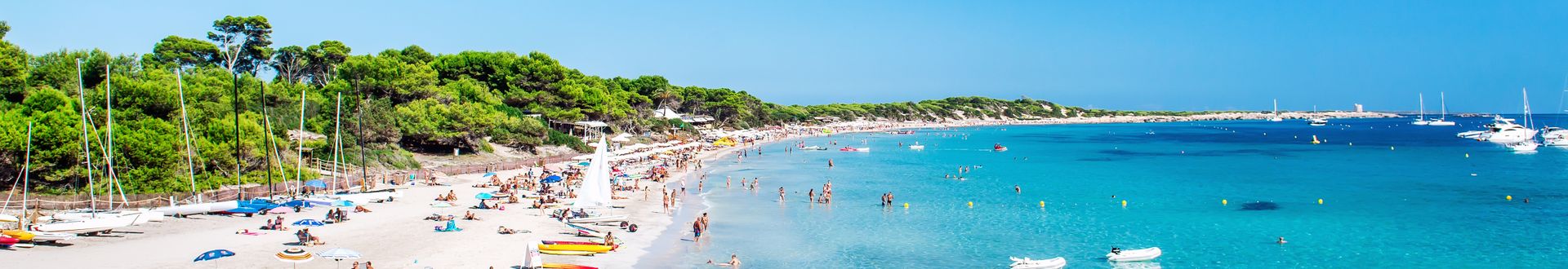 Weißer Sandstrand Ses Salines auf Ibiza mit türkisblauem Wasser, vielen Besuchern und Booten vor der Küste