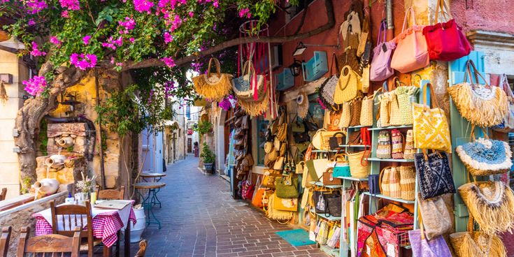 Enge Einkaufsstraße mit zahlreichen bunten Taschen und Körben aus Naturmaterialien, die an einer Wand hängen, und blühendem Bougainvillea darüber.