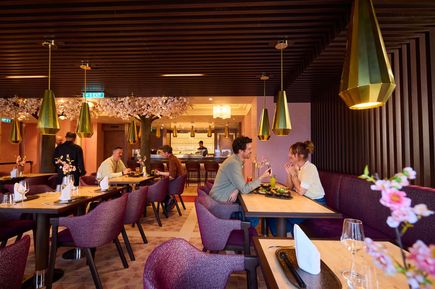 Modernes Restaurant mit warmem Licht und stilvoller Einrichtung: mehrere Gäste sitzen an Holztischen, trinken und essen; über ihnen hängen goldene, geometrische Pendelleuchten, im Hintergrund eine Bar sowie ein dekorativer Baum mit rosa Blüten.