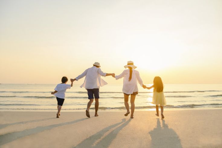 Familie geht Hand in Hand am Strand entlang in Richtung Sonnenuntergang, während sanfte Wellen an das Ufer rollen
