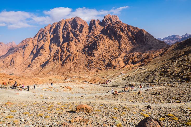 Der Berg Sinai in Ägypten erhebt sich mit seinen markanten Felsen, während Wanderer und Kamelreiter den Weg durch die karge Landschaft zurücklegen