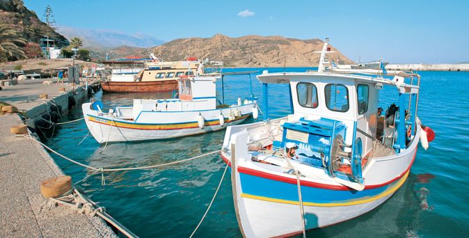 Fischerboote mit weißen Rümpfen und bunten Streifen liegen an einem Hafenpier vor einer felsigen Küstenlandschaft unter blauem Himmel.