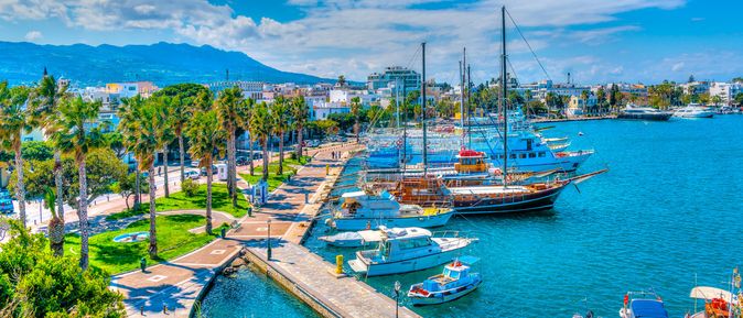 Hafenansicht in Kos, Griechenland, mit mehreren Booten im Wasser, Palmen entlang der Uferpromenade und einem klaren blauen Himmel.