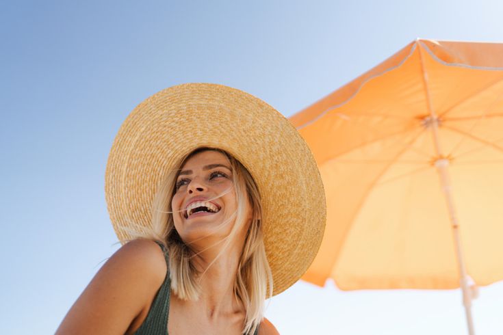 Frau mit großem, geflochtenem Strohhut sitzt unter einem orangefarbenen Sonnenschirm an einem sonnigen Tag