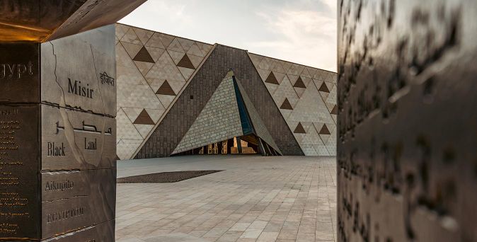 Blick durch eine Metallstruktur mit eingravierten Texten auf eine moderne Pyramidenarchitektur mit dreieckigen Mustern auf dem Platz