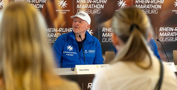 Joey Kelly sitzt an einem Tisch mit Mikrofon vor einer Wand mit dem Schriftzug ‚Rhein-Ruhr Marathon Duisburg‘ und trägt ein blaues Shirt mit dem Logo von ‚schausinsel reisen‘