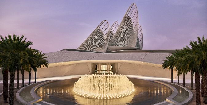 Beleuchteter Springbrunnen vor einem modernen Gebäude mit geschwungenen, netzartigen Dachstrukturen und Palmen an den Seiten