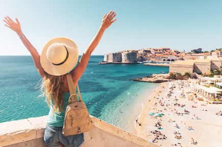 Person mit Sonnenhut blickt über den Banje-Strand und das türkisfarbene Meer auf die historische Altstadt von Dubrovnik