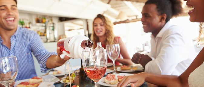 Mehrere Personen sitzen an einem gedeckten Tisch mit Tellern, Gläsern und verschiedenen Speisen, eine Person gießt ein Getränk in ein Weinglas.