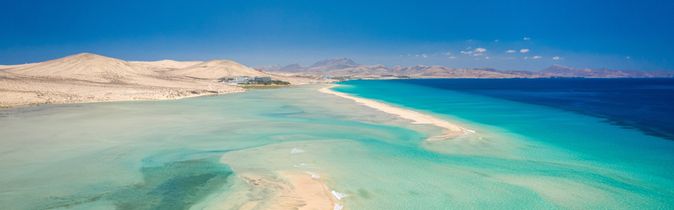 Luftaufnahme einer Sandbank im türkisfarbenen Meer vor einer kargen Küstenlandschaft unter klarem blauem Himmel