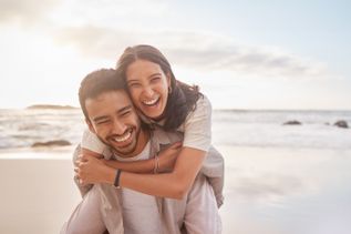 Mann trägt Frau auf dem Rücken am Strand, während beide sich umarmen und das weiche Licht des Sonnenuntergangs die Szene umgibt