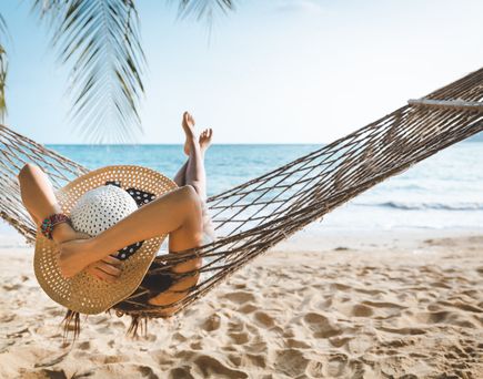 Frau mit Strohhut entspannt in einer Hängematte am Sandstrand mit Blick aufs Meer