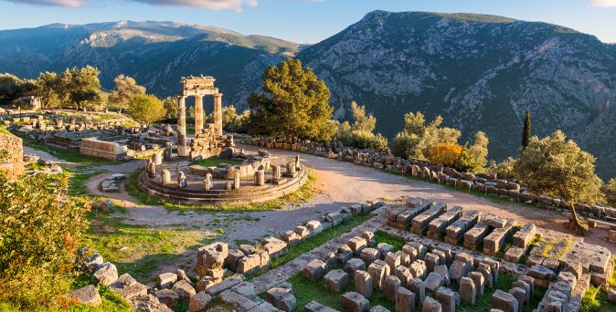 Antike Ruinen mit rundem Tempel und Säulen in Delphi, umgeben von Bergen und Bäumen bei Sonnenlicht.