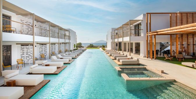 Zwei moderne Hotelgebäude mit Balkonen flankieren einen langen, klaren Pool. Liegen und Holzdecks sind entlang des Pools angeordnet.