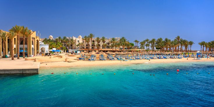 Strandpromenade einer ägyptischen Hotelanlage in Port Ghalib mit Palmen, Sonnenschirmen und Liegen am türkisblauen Meer unter klarem, blauem Himmel