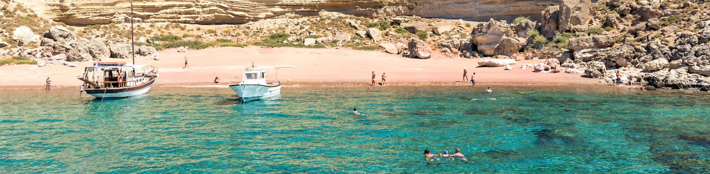 Roter Sandstrand auf Rhodos mit klarem türkisfarbenem Wasser, felsiger Küste und mehreren Menschen am Strand und im Wasser.