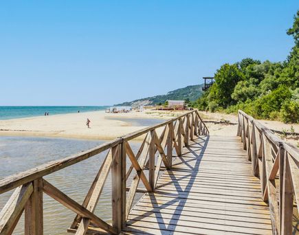 Holzsteg führt über Wasser zu einem Sandstrand in Albena, Bulgarien. Im Hintergrund sind Menschen am Strand und Bäume zu sehen.