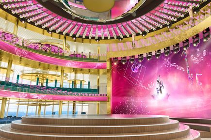 Theatrium der AIDAcosma mit großer Bühne, umlaufenden Sitzbereichen, goldenen und pinkfarbenen Designelementen sowie weitläufigem, modern beleuchtetem Innenraum