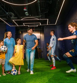 Mehrere Personen, darunter Kinder und Erwachsene, stehen auf grünem Kunstrasen in einem Raum mit dunklen Wänden und leuchtenden Deckenlampen, zwei lebensgroße Fußballspielerfiguren in Real-Madrid- und blauem Trikot.