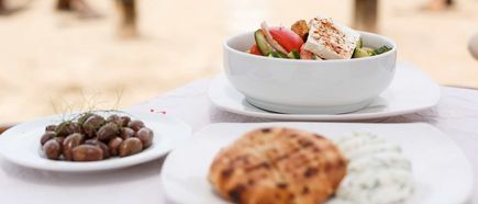 Weißer Teller mit griechischem Salat, daneben Teller mit Oliven und ein Teller mit Fladenbrot und Kräuterdip auf weißem Tischtuch.