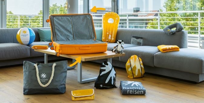 Offener orangefarbener Koffer auf einem Holztisch neben einem grauen Sofa mit diversen Reiseutensilien wie einem gelben und grauen Handtuch, einer Strandtasche, einem kleinen Stofftier und einem Wasserball.