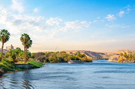 Boot auf dem Nil, Palmen am Ufer und sandige Hügel im Hintergrund unter blauem Himmel