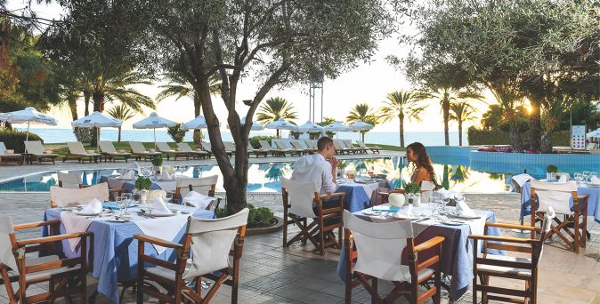 Zwei Personen sitzen an einem gedeckten Tisch mit weißen Stühlen und blauen Tischdecken neben einem Pool, umgeben von Bäumen und Sonnenschirmen.