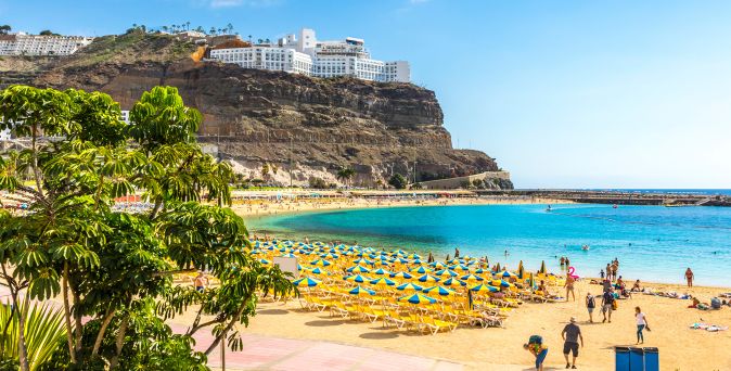Blick auf den Playa de Amadores auf Gran Canaria mit türkisblauer Badebucht, gelben Sonnenschirmen, Sandstrand und Hotels auf den Klippen oberhalb der Küste
