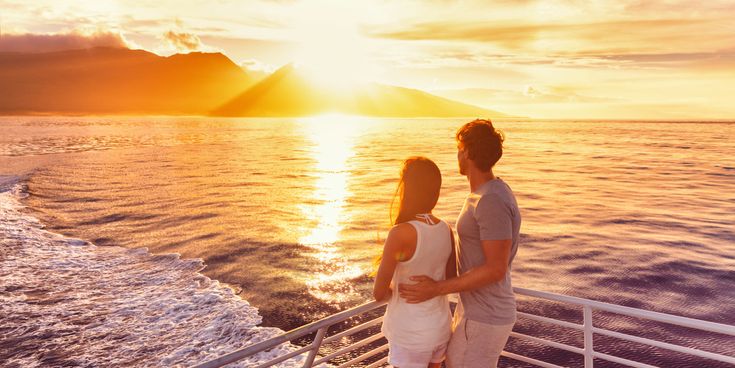 Zwei Personen stehen auf dem Außendeck eines Kreuzfahrtschiffes und blicken auf das Meer, während die Sonne hinter einer bergigen Küstenlandschaft untergeht und warmes Licht auf das Wasser fällt