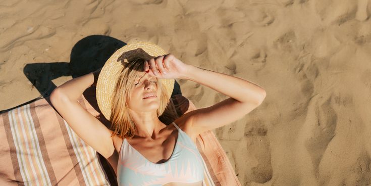 Person liegt entspannt auf einem gestreiften Handtuch im warmen Sand und hält einen Strohhut über dem Gesicht, während die Sonne die Strandfläche beleuchtet