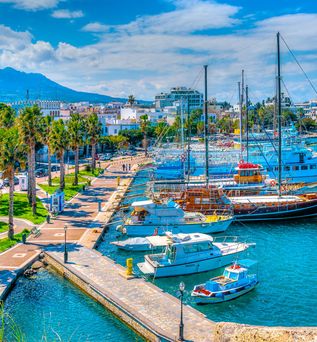 Hafenansicht in Kos, Griechenland, mit mehreren Booten im Wasser, Palmen entlang der Uferpromenade und einem klaren blauen Himmel.