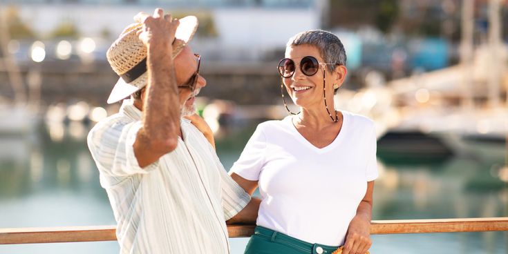 Mann und Frau stehen auf dem Außendeck eines Kreuzfahrtschiffs an einer Reling, mit sonnigem Hafenpanorama im Hintergrund