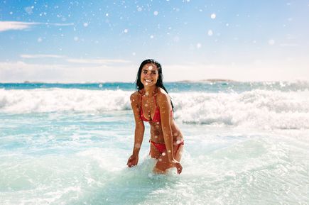 Lächelnde Frau in rotem Bikini steht im flachen Wasser, während Wellen brechen und Wassertröpfchen in der Luft glitzern