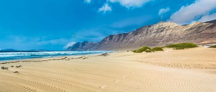 Playa de Famara auf Lanzarote mit weitläufigem Sandstrand, Dünen im Vordergrund, Atlantikbrandung und dem steil aufragenden Famara-Massiv unter blauem Himmel
