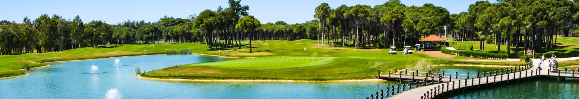 Panorama eines Golfplatzes in Belek, Türkei, mit breitem Wasserhindernis, Springbrunnen, Holzsteg und grünen Fairways, umgeben von dichtem Wald und sonnigem Himmel