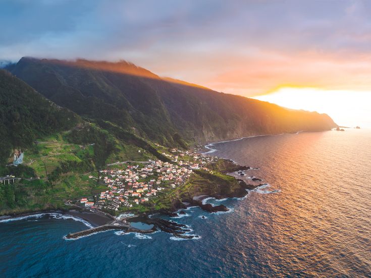Luftaufnahme von Seixal auf Madeira bei Sonnenuntergang mit Küstenort, terrassenförmig angelegten Hängen, steilen grünen Bergen und dem Atlantik entlang der zerklüfteten Küste