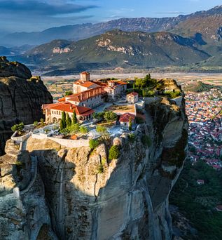 Kloster auf hohem Felsen mit roten Dächern, umgeben von Bergen und einer Stadt im Tal.