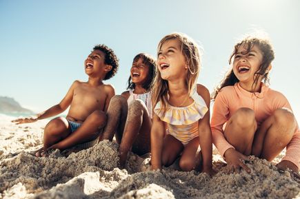 Gruppe fröhlicher Kinder sitzt im Sand am Strand und lacht während eines sonnigen Badetag-Urlaubs