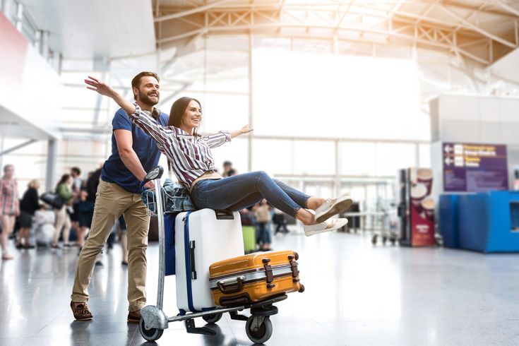 Mann schiebt Frau mit ausgestreckten Armen auf Gepäckwagen mit zwei Koffern in Flughafenhalle