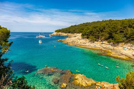 Felsige Badebucht bei Pula in Istrien, Kroatien, mit kristallklarem Wasser, Badenden und Booten vor grüner Küstenlandschaft an der Adria