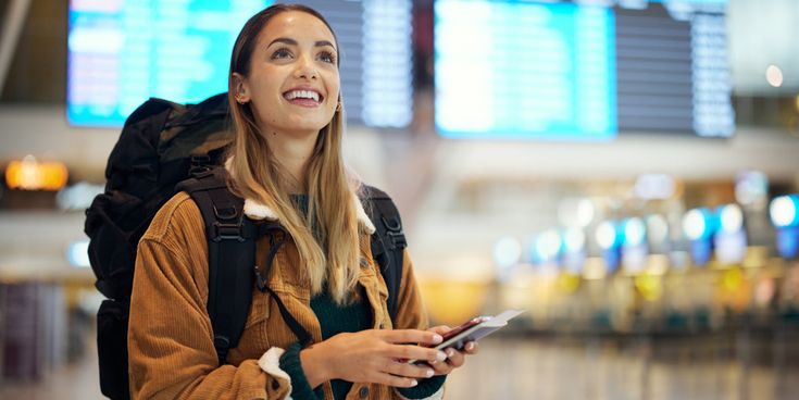 Junge Frau mit Rucksack, Smartphone und Reisepass lächelt in einer Flughafenhalle vor den großen Anzeigetafeln